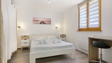 A hotel room with a bed, bedside tables, a desk, a chair, a radiator, and a window with blinds.