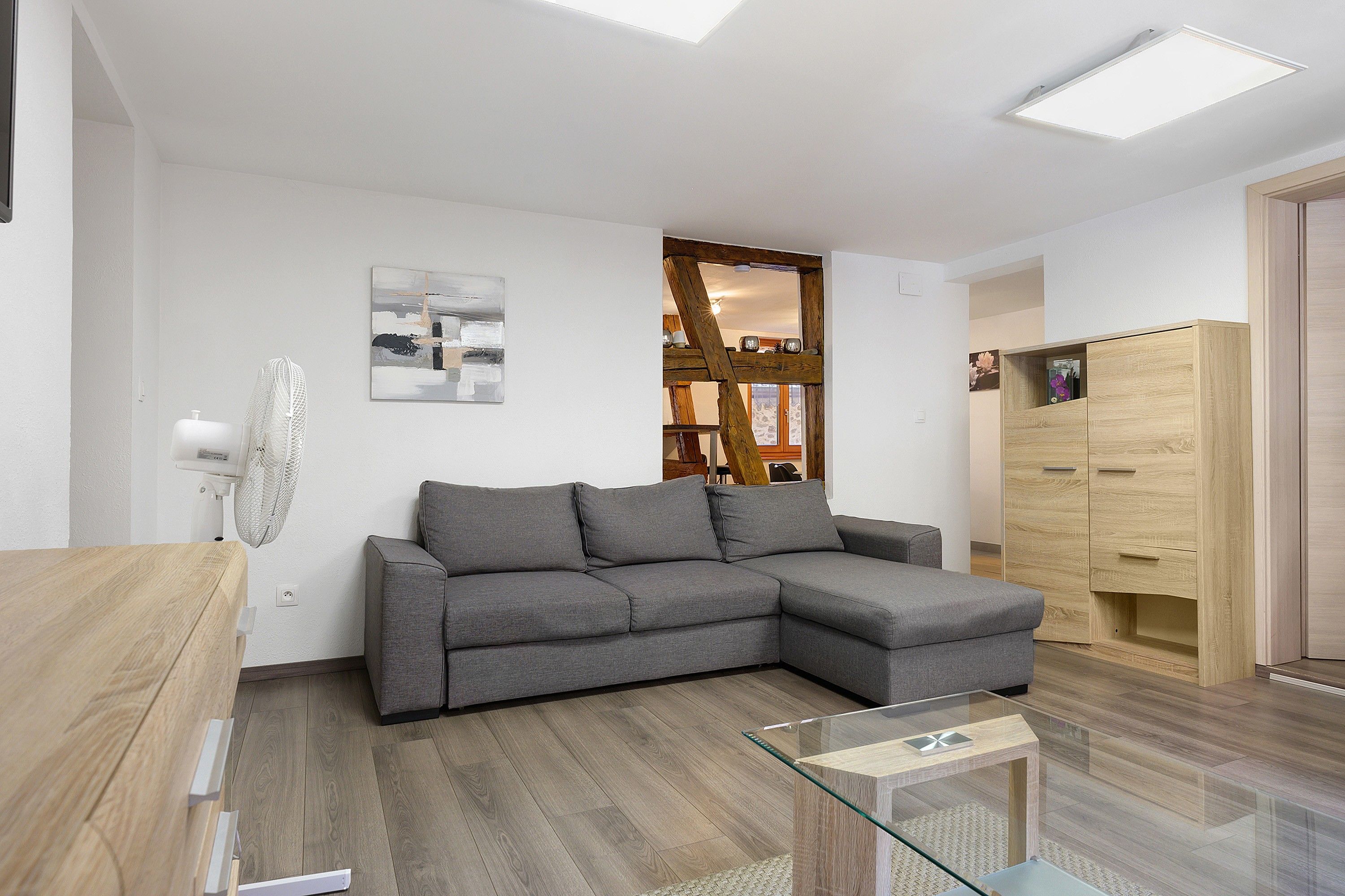 A modern living room with a grey sectional sofa, a glass coffee table, wooden flooring, a white wall with a painting, and a wooden cabinet.