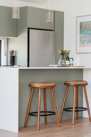 A kitchen with a stainless steel refrigerator, two wooden stools with black metal bases, and hanging pendant lights. - 'Eclipse' at Small Batch (Budgee Budgee)