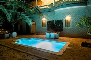 A swimming pool with a tiled deck, a palm tree, and a building with a textured facade and two lit windows. - Dar Rouissi (Tozeur)