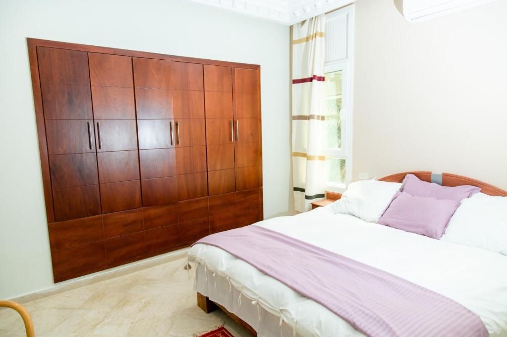 Een slaapkamer met een grote kledingkast, een bed met wit beddengoed en een paarse plaid, en een raam met gordijnen.