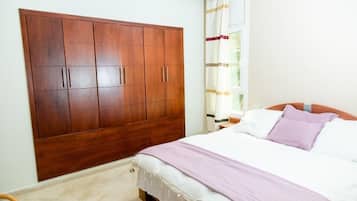 Een slaapkamer met een grote kledingkast, een bed met wit beddengoed en een paarse plaid, en een raam met gordijnen.