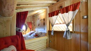 Interior de una cabaña con una cama, una silla roja y una ventana con cortinas.