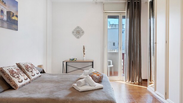 Een slaapkamer met een bed, kussens, een nachtkastje, een vaas en uitzicht op buiten.