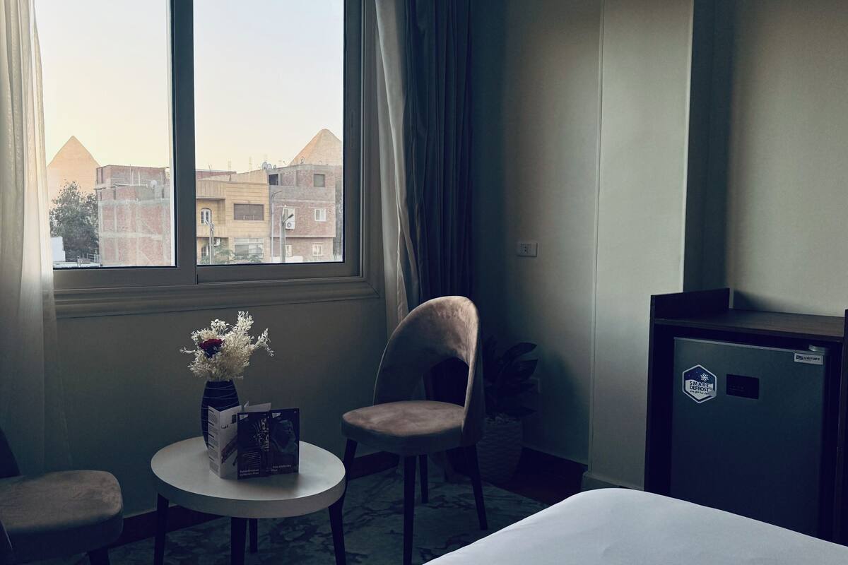 A hotel room with a bed, two chairs, a small table with a vase and a box, a window with curtains, and a view of buildings outside.