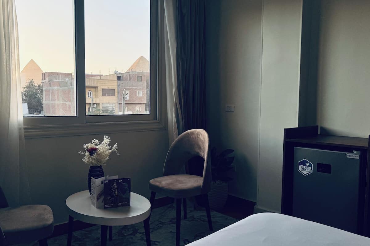 Hotellihuone, jossa on sänky, kaksi tuolia, pieni pöytä, maljakko ja rasia, ikkuna verhoilla sekä näkymä ulkona oleviin rakennuksiin.