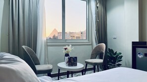Habitación de hotel con cama, dos sillas, una mesita con un florero y un libro, una planta y vistas a una pirámide a través de la ventana.