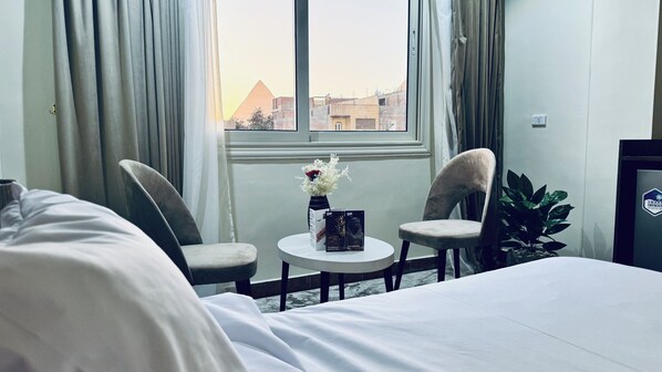Habitació d'hotel amb llit, dues cadires, taula petita amb un ram i un llibre, finestra amb vista a edificis i una planta.