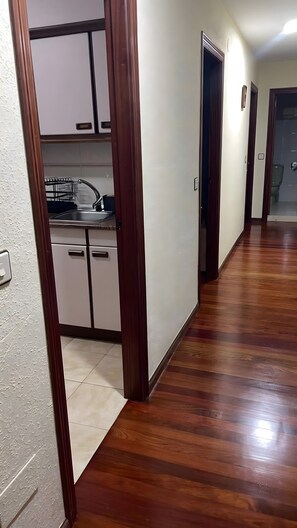 Un couloir avec un plancher en bois, un coin cuisine avec des placards, un évier et des toilettes visibles à travers une porte ouverte.