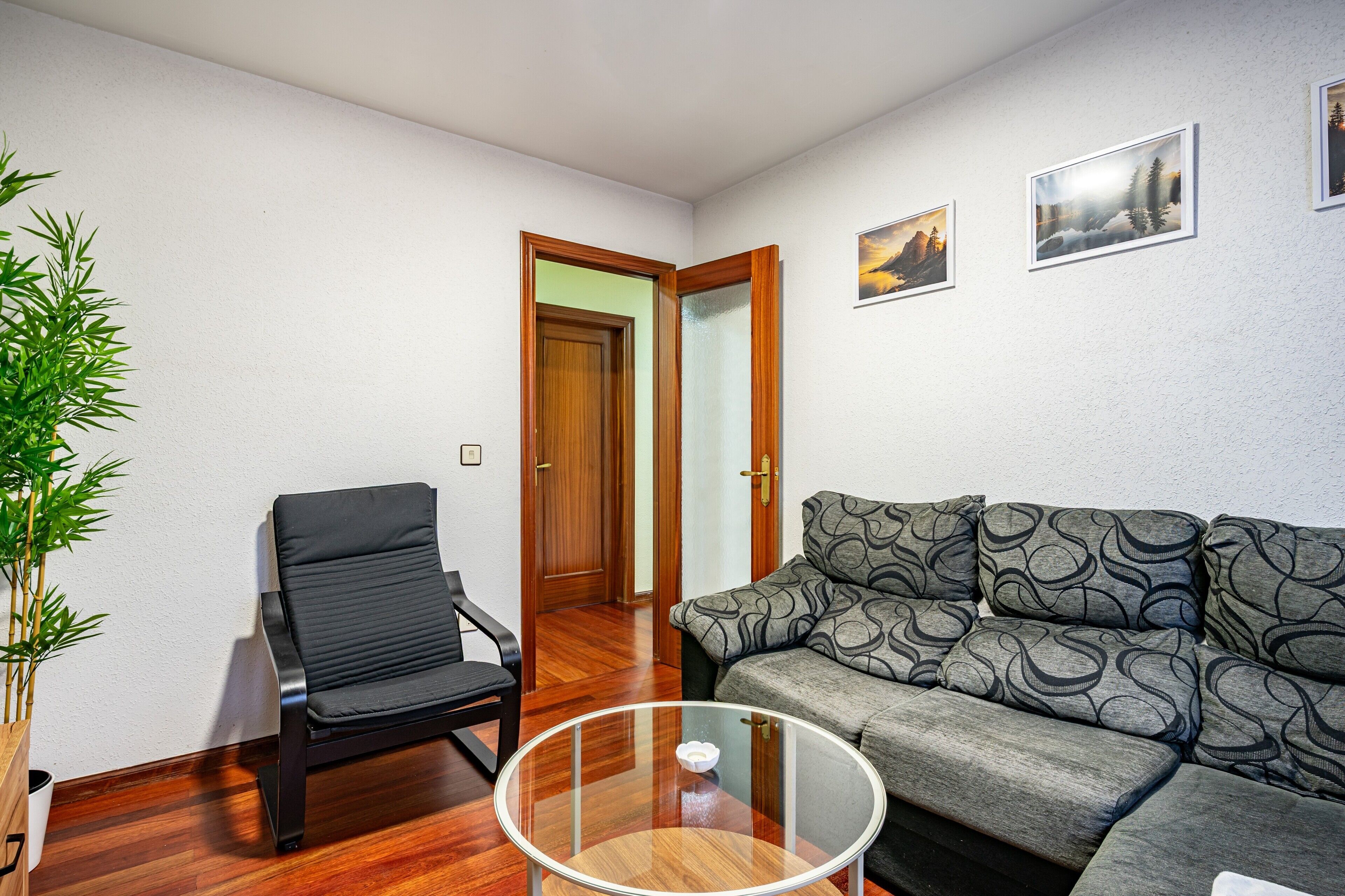 Una habitación con suelo de madera, una silla negra, un sofá gris con estampado, una mesa de centro de cristal y una puerta de madera.