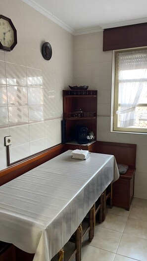 Une pièce avec une longue table, une horloge, une étagère remplie d’assiettes et une fenêtre avec des rideaux.
