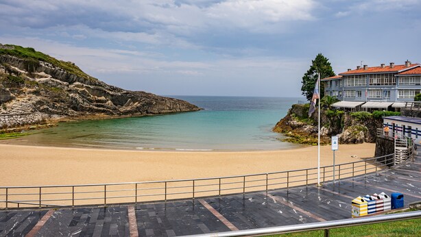 A beach with a rocky cliff, a building with a red roof, and a paved walkway. - Apartment 'Centro Y Mar En Llanes' with Sea View and Wi-Fi (Llanes)