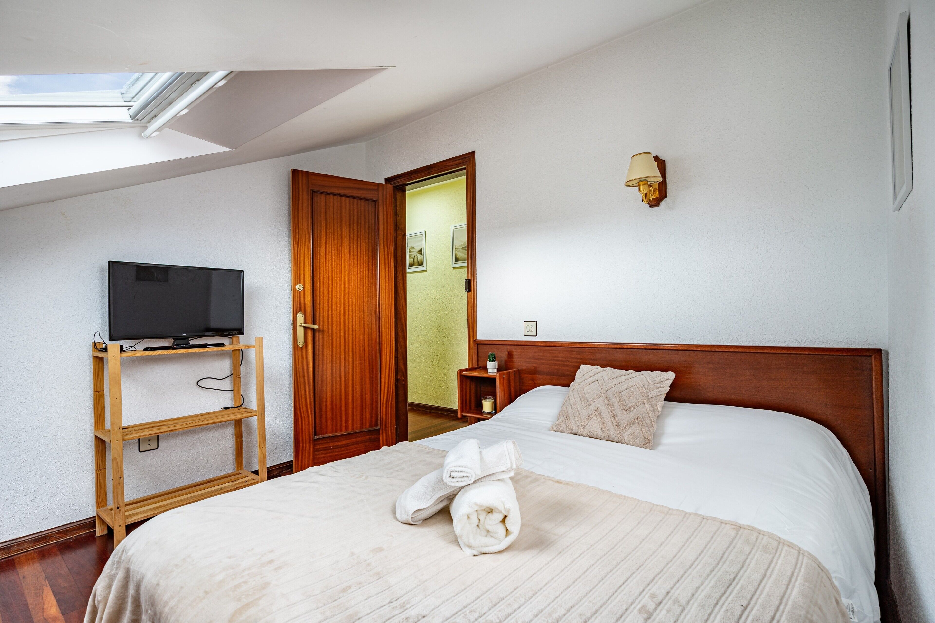 Habitación de hotel con una cama, un televisor sobre un soporte de madera, una mesita de noche con una lámpara y una puerta que da a otra habitación.
