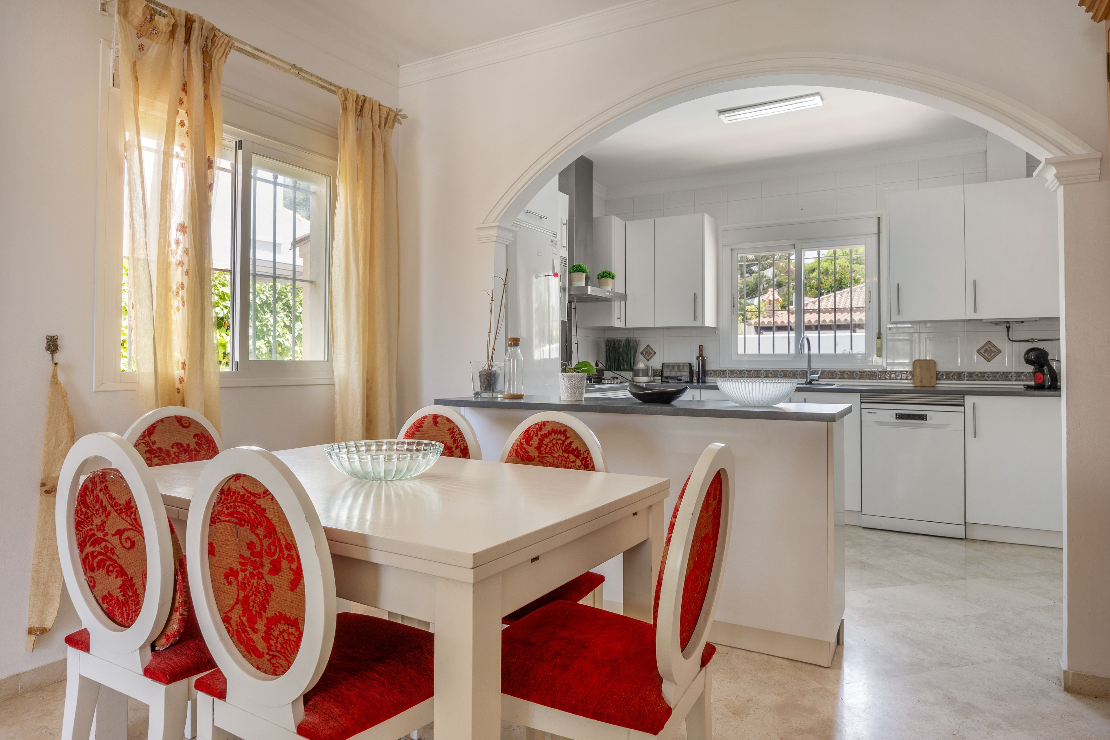 Un comedor con una mesa blanca y sillas tapizadas rojas, una cocina con muebles blancos, y una vista a través de un arco.
