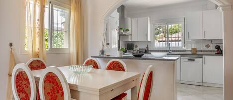 Un comedor con una mesa blanca y sillas tapizadas rojas, una cocina con muebles blancos, y una vista a través de un arco.