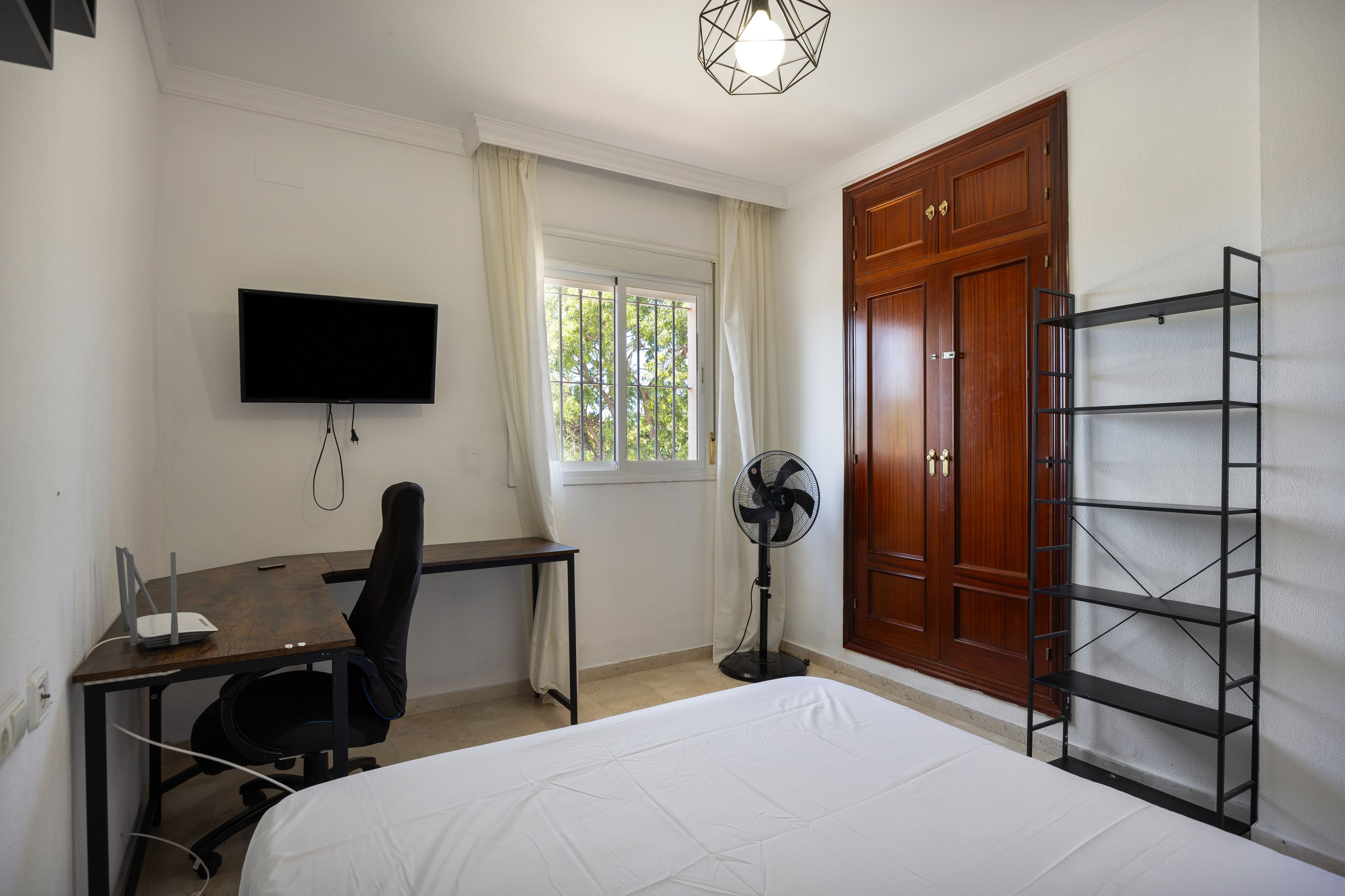 Una habitación con cama, escritorio, silla, televisor, ventana, ventilador, armario y estanterías.