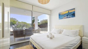 Un dormitorio con cama, un cuadro, una silla de mimbre y vistas a través de un ventanal.