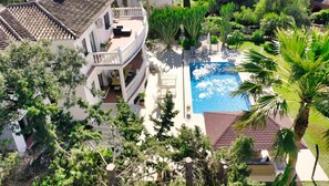 Ein Pool, umgeben von üppiger Vegetation, ein weißes Gebäude mit Balkon und ein Ziegeldach.