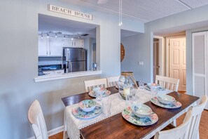 A dining area with a wooden table, white chairs, and a kitchen visible through an open window. - Ocean and racquet Club: steps to ocean, pool, tennis and pickleball (saint augustine)