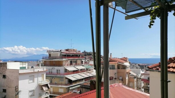 Um edifício de vários andares com varandas, telhado vermelho e vista para o mar e montanhas ao fundo.