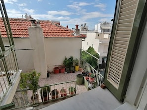 A balcony with potted plants, a metal railing, and a view of neighboring buildings with red-tiled roofs.