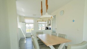 Uma sala de jantar com mesa de madeira e cadeiras brancas, uma cozinha ao fundo e vista para o exterior.
