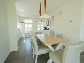A dining area with a wooden table and white chairs, a kitchen in the background, and a view of the outside. - Florijn Sauna 6 (Uitdam)