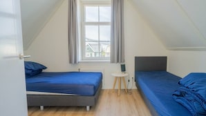 Um quarto de sótão pequeno e triangular com duas camas, uma janela com cortinas e uma mesinha com luminária.