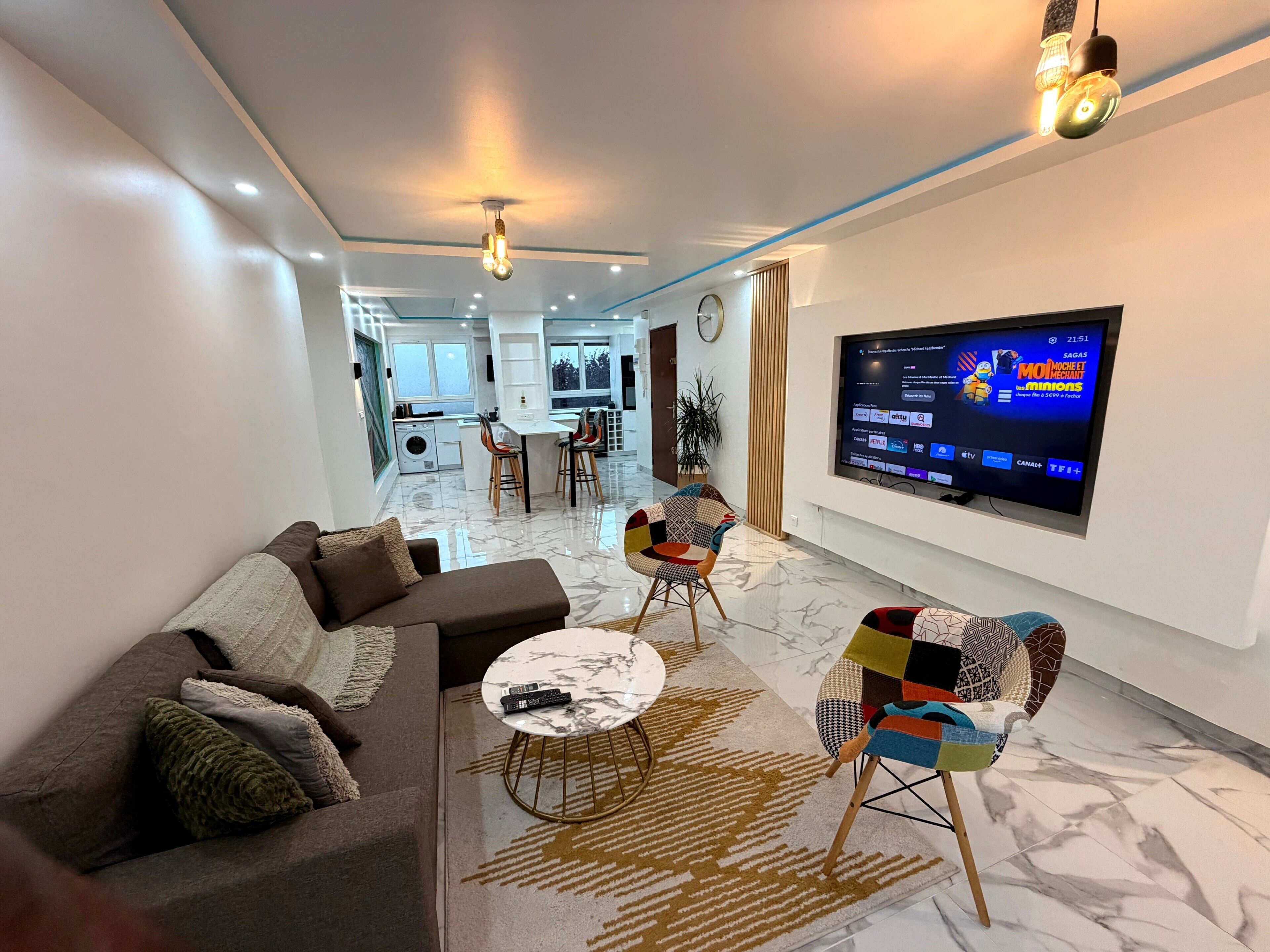 A modern living room with a sofa, coffee table, and colorful chairs, a TV mounted on the wall, and a kitchen in the background.