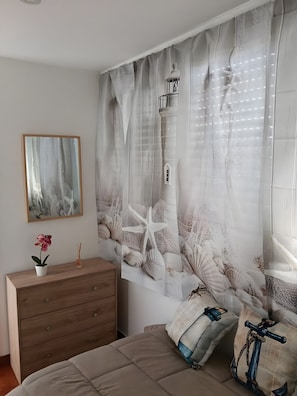 Een slaapkamer met een bed, een houten dressoir, een lijst met afbeelding en een decoratieve wand met een vuurtoren en schelpen.