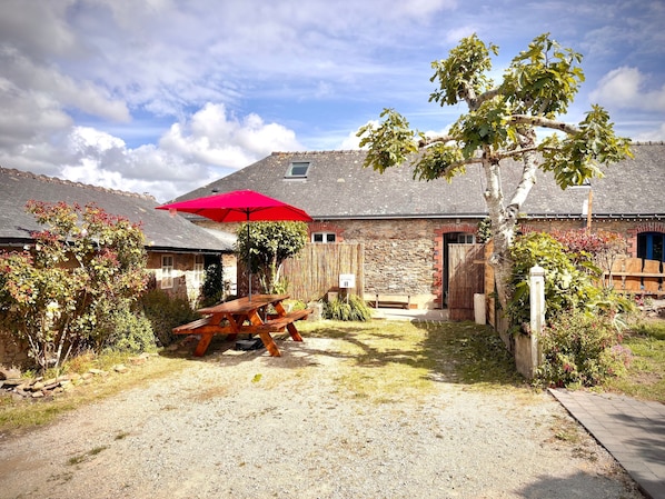 A rustic stone building with a wooden fence, a red umbrella, and a picnic table in the yard.