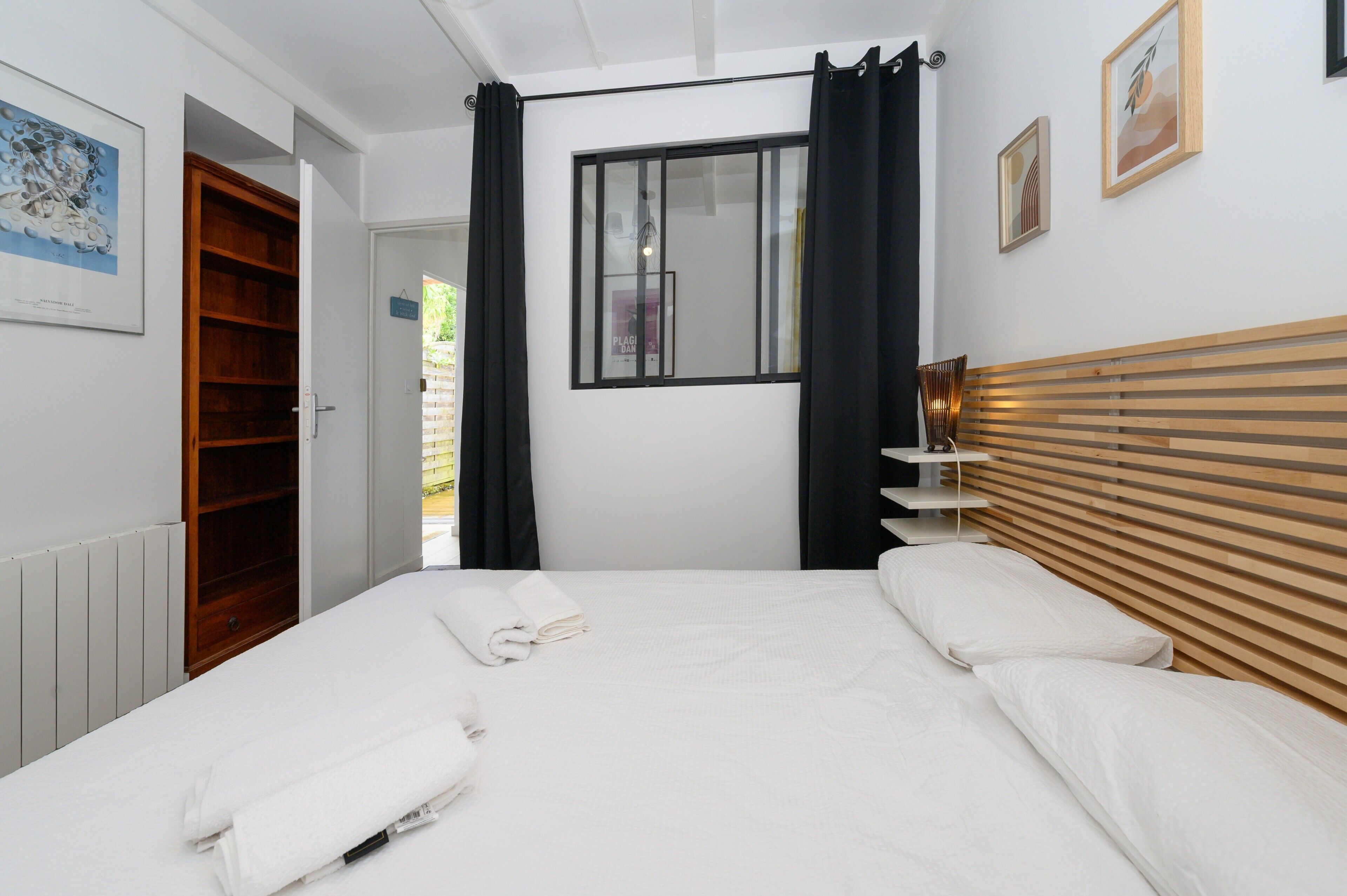 A bedroom with a bed, a wooden headboard, a window with curtains, a bedside table, and a wall-mounted shelf.