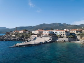 Un pueblo costero con edificios a lo largo del paseo marítimo, un muelle y montañas al fondo.