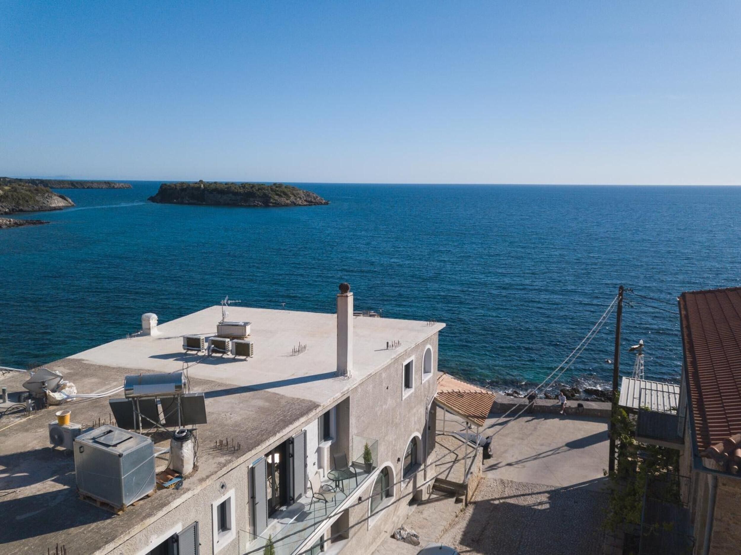 A coastal building with a rooftop terrace overlooking the sea, a small island in the distance, and a clear sky.