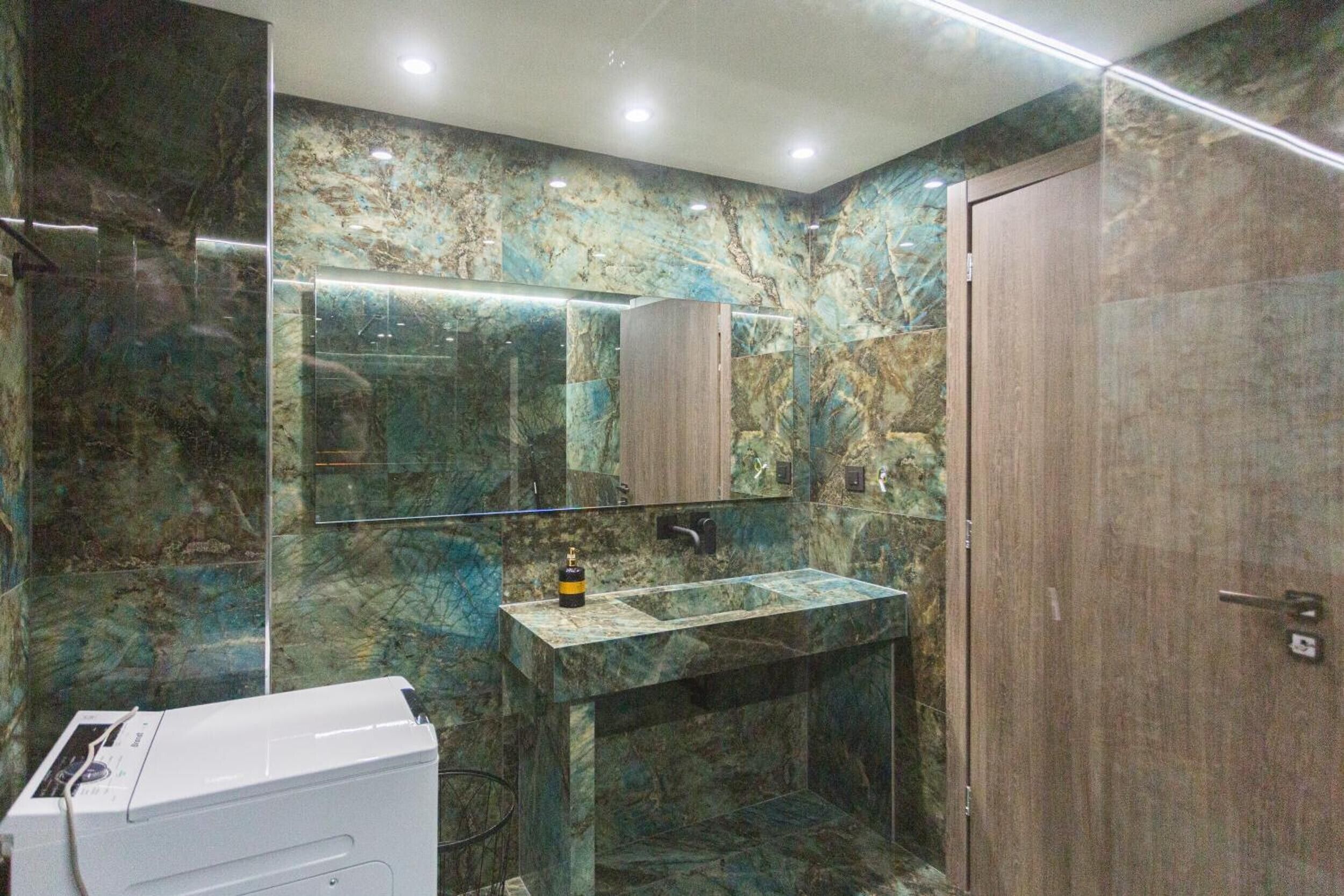 A bathroom with a marble wall, a sink, a mirror, and a door.