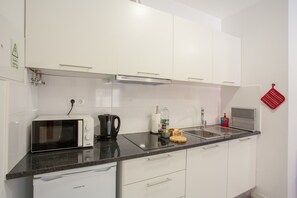 Een keuken met witte kastjes, een magnetron, een waterkoker, een spoelbak en een rode handdoek aan de muur.