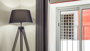 Una lámpara de pie con trípode, una ventana con barrotes metálicos y una puerta con detalle rojo.