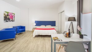 Habitación de hotel con una cama, un escritorio, dos sillas azules, una lámpara y un cuadro en la pared.