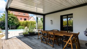 En overdækket terrasse med et træbord og stole, en pool og udsigt til en rød bygning.