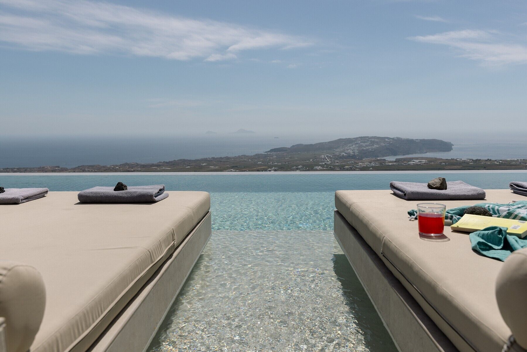 On aperçoit clairement une piscine, des chaises longues et des serviettes, un verre d’une boisson rouge, et, au loin, une côte.