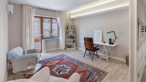 A modern living room with a glass coffee table, a white sofa, a patterned rug, a bookshelf, and a desk with a chair.