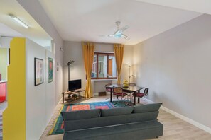 Ein Wohnzimmer mit einer Couch, einem Esstisch, Stühlen, einem Fernseher, einem Deckenventilator und einem Fenster mit Vorhängen.