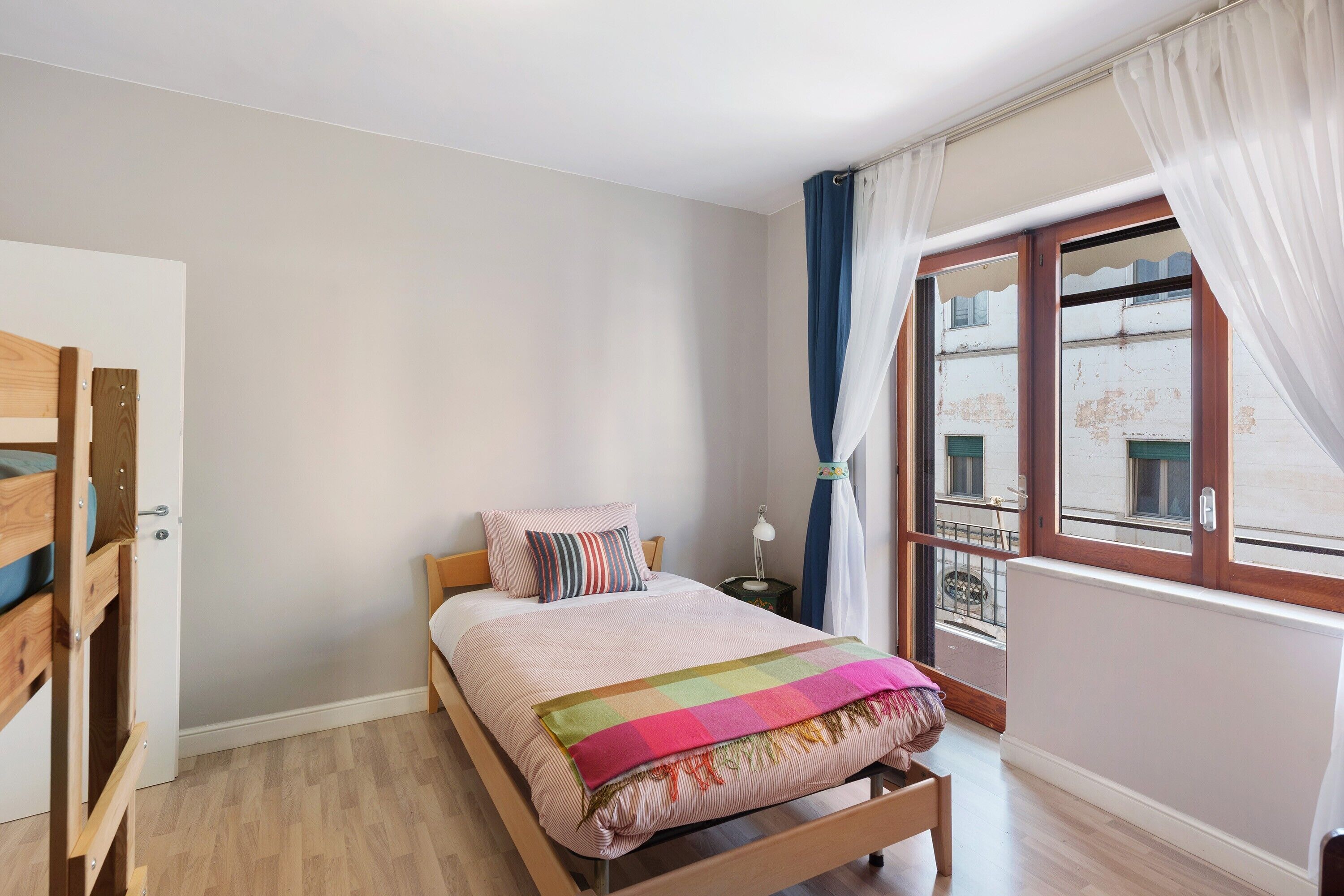 A bedroom with a bed, a nightstand, a window with curtains, and a view of buildings outside.
