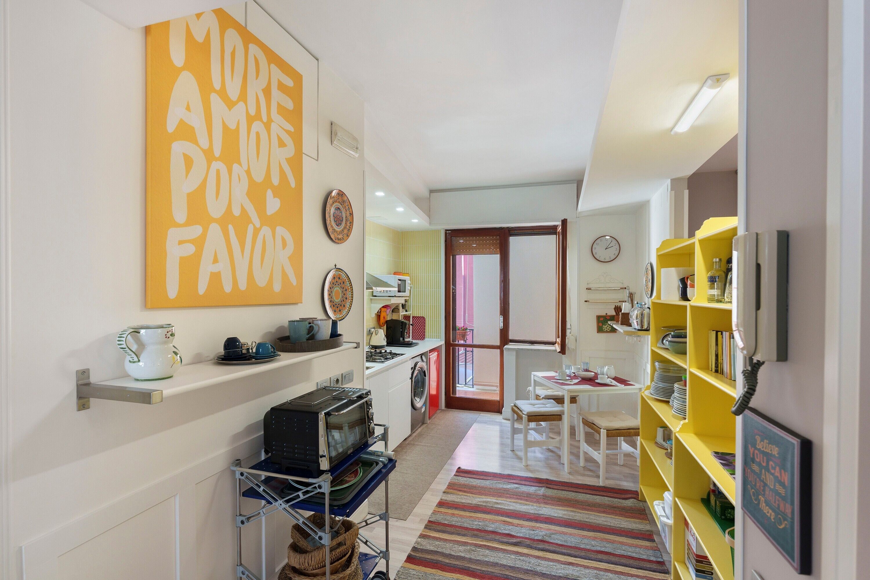 A compact kitchen with a microwave, stove, and sink, a dining area with a table and chairs, and a yellow bookshelf.