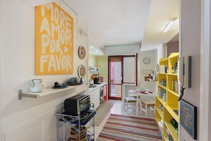 Een compacte keuken met een magnetron, fornuis en wastafel, een eethoek met een tafel en stoelen, en een geel boekenrek.