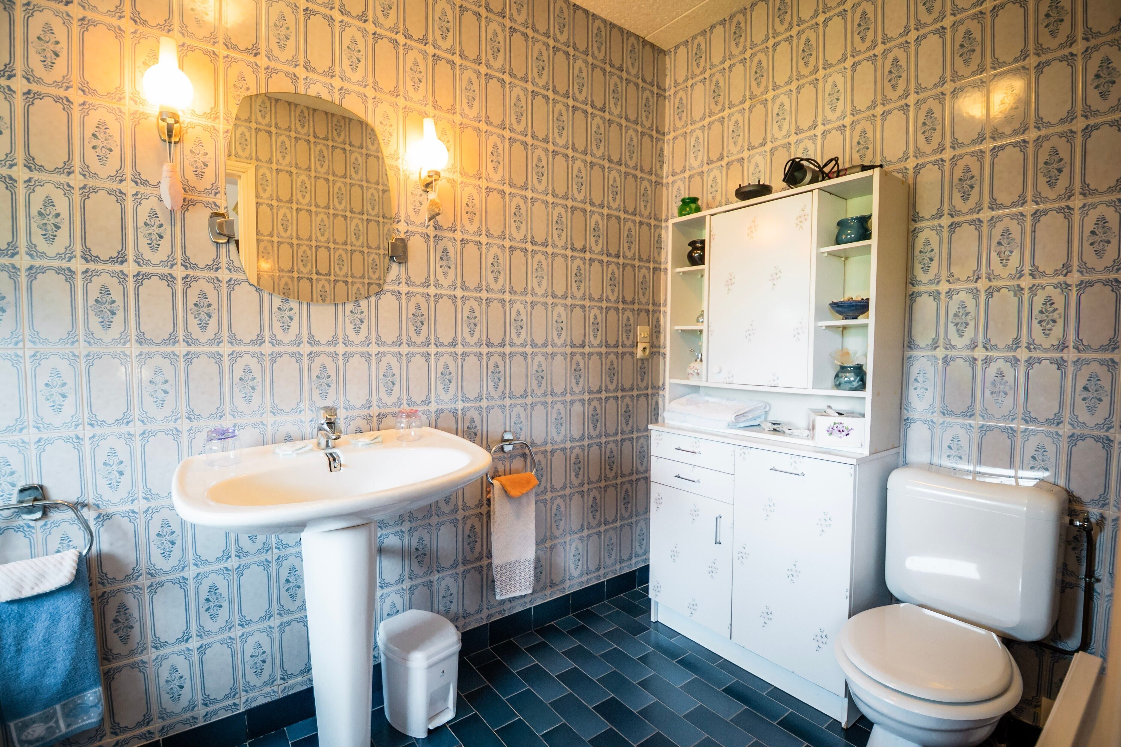 Ein Badezimmer mit weißem Waschbecken, Toilette und Spiegel. Ein Ablagefach mit Pflegeprodukten und ein Handtuch hängen an der Wand.