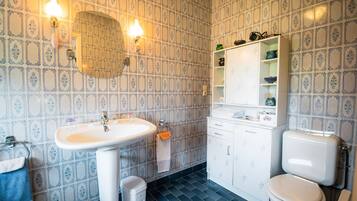 Ein Badezimmer mit weißem Waschbecken, Toilette und Spiegel. Ein Ablagefach mit Pflegeprodukten und ein Handtuch hängen an der Wand.
