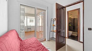 A room with a red sofa, a wooden door, a glass door leading to a balcony, and a small shelf with decorative items.