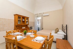Une pièce comprenant une table à manger dressée pour deux personnes, un buffet en bois, un lit recouvert d’une couette blanche et une fenêtre avec un rideau.