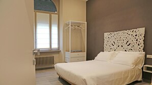 Une chambre à coucher avec un lit, une fenêtre avec des stores, une table de chevet et une étagère fixée au mur.
